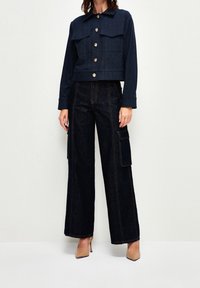 Chaqueta de mezclilla con dos bolsillos en el pecho, cierre de botones y un diseño recortado en azul oscuro, combinada con pantalones cargo de pierna ancha en azul oscuro.