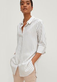 Camicia bianca con bottoni, caratterizzata da righe dorate verticali, maniche arrotolate e tasche laterali. Tessuto leggero con vestibilità rilassata e orlo curvo.