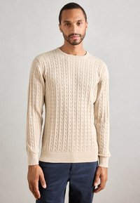 Άνδρας με κοντά μαλλιά και μούσι, ντυμένος με μπεζ πλεκτή μπλούζα και σκούρα παντελόνια, στέκεται μπροστά σε απλή ανοιχτή γκρίζα φόντο.