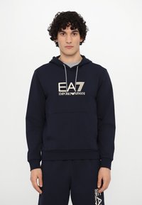 Navy blue hoodie with a kangaroo pocket, featuring a light beige logo "EA7 EMPORIO ARMANI" at the front and drawstrings on the hood.
