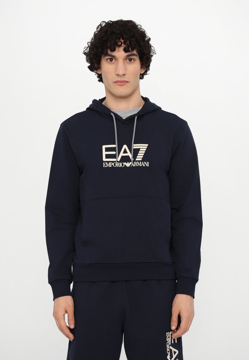 Navy blue hoodie with a kangaroo pocket, featuring a light beige logo "EA7 EMPORIO ARMANI" at the front and drawstrings on the hood.