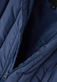 Mørkeblå isoleret jakke indvendig med glat tekstur, der har en fremtrædende lynlås og syede detaljer langs kanterne og lommeåbningerne.