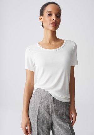 Vrouw draagt een wit kortemouwen topje met ronde hals en een grijze, hooggetailleerde gestructureerde broek, staand voor een effen lichte achtergrond.