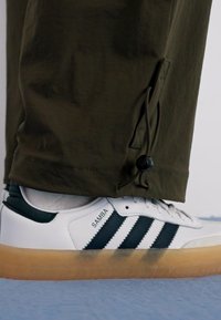 White Adidas Samba sneakers with navy stripes and gum sole, paired with olive green pants featuring adjustable cuffs and a smooth texture.