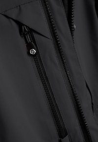 Melnas jakas auduma tuvplāns ar rāvējslēdzēja aizdari, kas aprīkota ar teksturētu vilkšanas cilpu un izšūtiem akcentiem, atklājot gludas un matētas faktūras.