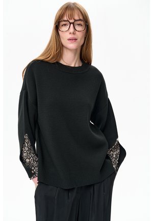 Ciemnozielony sweter z okrągłym dekoltem, luźnym krojem, ozdobnym kwiatowym wzorem na rękawach, prążkowanym dołem oraz dużymi czarnymi spodniami.