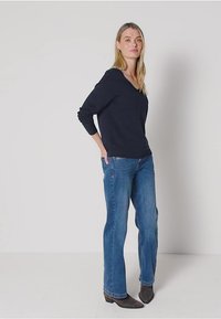 T-paita syvällä V-pääntiellä, tummansininen pitkähihaisuus, yhdistettynä vaaleansinisiin suoriin farkkuihin ja mustiin nilkkabootseihin, yksinkertaista valkoista taustaa vasten.