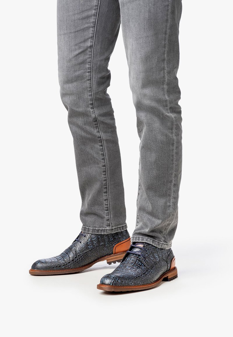 Zapatos de cuero con patrón de cocodrilo azul y acentos marrones, combinados con jeans ajustados grises, que exhiben una textura detallada y un diseño elegante.