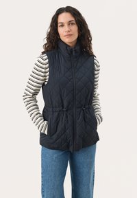 Gestepptes marineblaues Weste mit hohem Kragen, Reißverschluss vorne und Kordelzug in der Taille; getragen über einem gestreiften Langarmoberteil und kombiniert mit blauen Jeans.