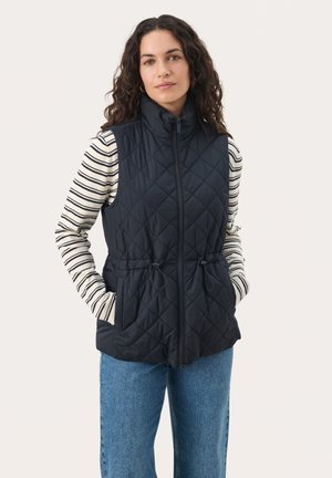 Quilted navy vest with high collar, zip front, and drawstring waist; worn over a striped long-sleeve top and paired with blue jeans.