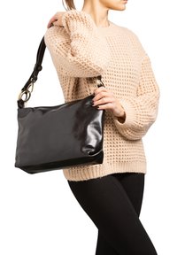 Zwarte leren handtas met een glanzende afwerking, rechthoekige vorm en een afneembare schouderband met gouden details. Wordt onder de arm gedragen door een model.