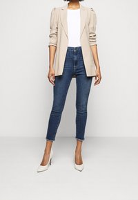 Blazer beige con mangas abullonadas, camisa blanca ajustada, jeans azul de cintura alta y tacones blancos con punta, de pie sobre un fondo neutro.