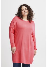 Maglione corallo oversize con maniche lunghe e scollatura rotonda, caratterizzato da una texture morbida e spacchi laterali, abbinato a jeans scuri.