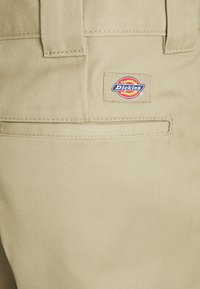 Beige Stoffhose mit einer Gesäßtasche, die mit einem blauen und roten Dickies-Logoschild versehen ist, sichtbaren Nähten und einer glatten Textur.