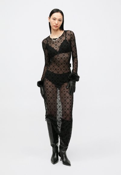 Čierne priehľadné šaty s kvetinovým vzorom, dlhými rukávmi a bočnými rozparkami, doplnené čiernymi čižmami nad kolená a čiernymi rukavicami s kožušinovými akcentmi.