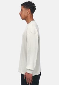 Camisa de manga larga de canalé gris claro con cuello redondo y mangas acampanadas, exhibida en un modelo de lado, mostrando una textura suave.