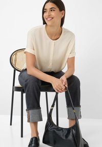 Beige blouse with button placket, rolled sleeves; dark jeans with cuff accents; black leather bag; model seated on a cane-backed chair.