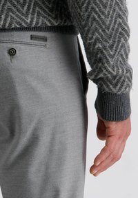 Grau gemusterter Pullover mit Zickzacktextur, kombiniert mit hellgrauen Wollhosen, die einen Knopfverschluss und ein Etikett an der Taille haben.