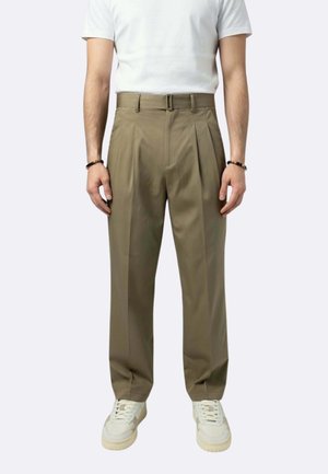 Uomo che indossa pantaloni oliva a vita alta e plissettati con cintura abbinata, sneakers bianchi, camicia bianca a maniche corte e braccialetti su entrambi i polsi.