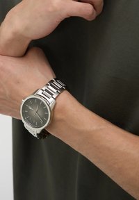 Srebrna ura iz nerjavečega jekla z okroglo površino, črnim številčnikom, srebrnimi kazalci in povezanimi kovinskimi zapestnicami, ki jo nosimo na zapestju.