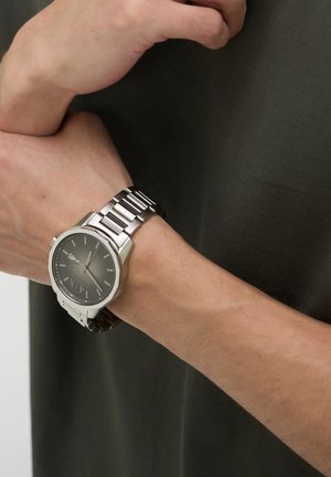 Srebrna ura iz nerjavečega jekla z okroglo površino, črnim številčnikom, srebrnimi kazalci in povezanimi kovinskimi zapestnicami, ki jo nosimo na zapestju.