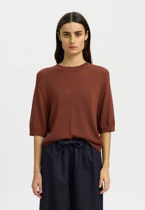 Femme aux longs cheveux foncés portant un pull à manches courtes couleur rouille et un pantalon sombre à taille élastique avec un cordon noué.