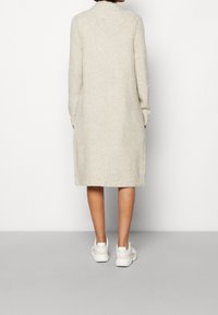 Knielanges, beiges Strickkleid mit langen Ärmeln, gerippter Textur und hohem Kragen, kombiniert mit weißen Sneakers.