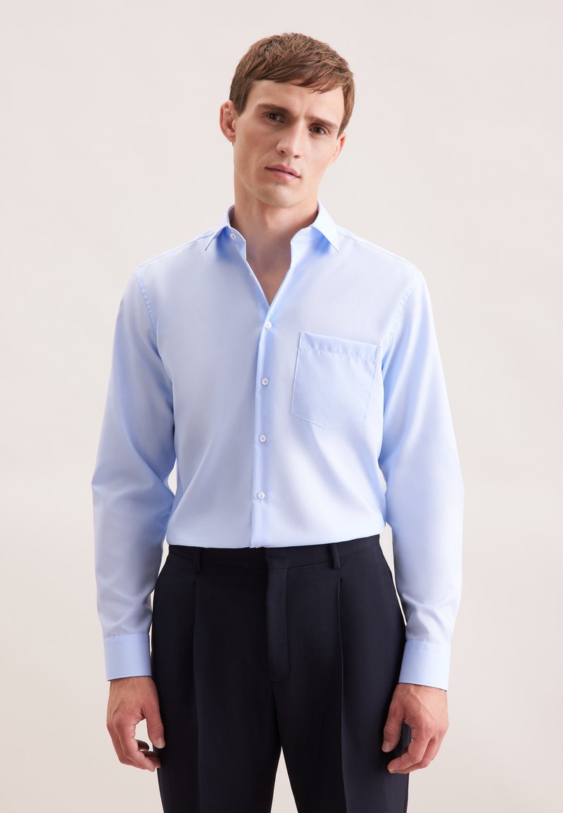 Camicia a maniche lunghe blu chiaro con colletto button-down e una tasca sul petto. Il tessuto sembra liscio e leggero. Abbinata a pantaloni scuri.