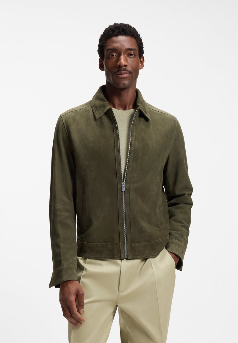 Homme portant une veste en daim vert olive sur une chemise vert clair et un pantalon beige, debout devant un fond blanc.