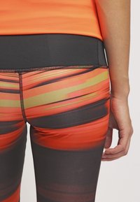Leggings taille haute en orange vif et noir, présentant un motif rayé et un tissu lisse et extensible. Coutures détaillées le long des coutures.