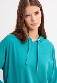 Sudadera turquesa con un corte holgado, capucha con cordón y textura suave. Presenta un bolsillo canguro y detalles acanalados en los puños.