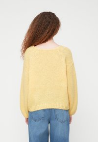 Πλεκτό κίτρινο πουλόβερ με άνετη γραμμή, μανίκια με ριμπ μανσέτες και φαρδιά λαιμόκοψη, συνδυασμένο με μπλε τζιν παντελόνι.