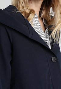 Persona rubia que lleva un abrigo azul marino oscuro con capucha sobre una camisa blanca con rayas verticales negras.