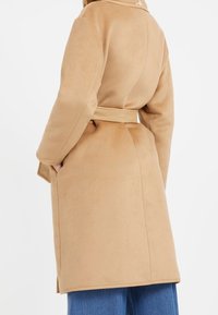 Lange tan kleurige jas van zacht stof, met een brede kraag, een ceintuur in de taille en zijzakken. Gladde textuur met subtiele stiksels.