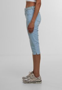 Jeans capri azzurri chiari con un design aderente, vita media e spacchi alla caviglia. Abbinati a scarpe da corsa beige con accenti grigi.