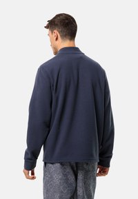 Giacca in pile blu marina con colletto, caratterizzata da una texture morbida e maniche lunghe. Abbinata a pantaloni grigi con motivo, che mostrano un fit rilassato.