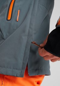 Main tenant une fermeture éclair latérale et une tirette en cuir sur une veste grise avec une fermeture éclair orange, portée par-dessus un pantalon orange.