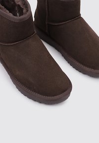 Botines de ante marrón con un suave forro de piel, puntera redondeada y suela de goma. El diseño incluye una banda cosida en la parte superior.