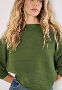 Maglione verde con texture, collo ampio e vestibilità morbida, con dettagli a coste sulle maniche. Abbinato a jeans in denim blu.