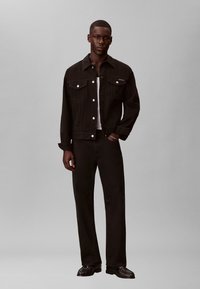 Hombre con chaqueta y pantalones de mezclilla negros y camisa blanca, de pie con una mano en el bolsillo sobre un fondo gris liso.