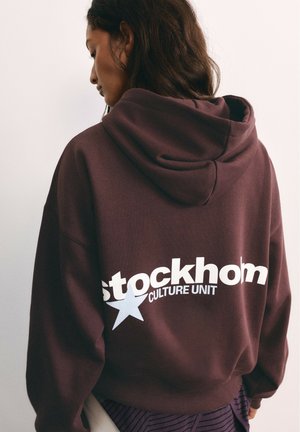 Femme aux cheveux foncés portant un sweat à capuche bordeaux avec "Stockholm Culture Unit" et une étoile imprimés dans le dos.