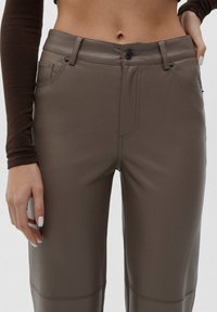 Braune Kunstlederhosen zeichnen sich durch ihr figurbetontes Design, Fronttaschen und dezente Stickdetails aus. Der Bund ist mit einem Knopfverschluss und Schlaufen ausgestattet.