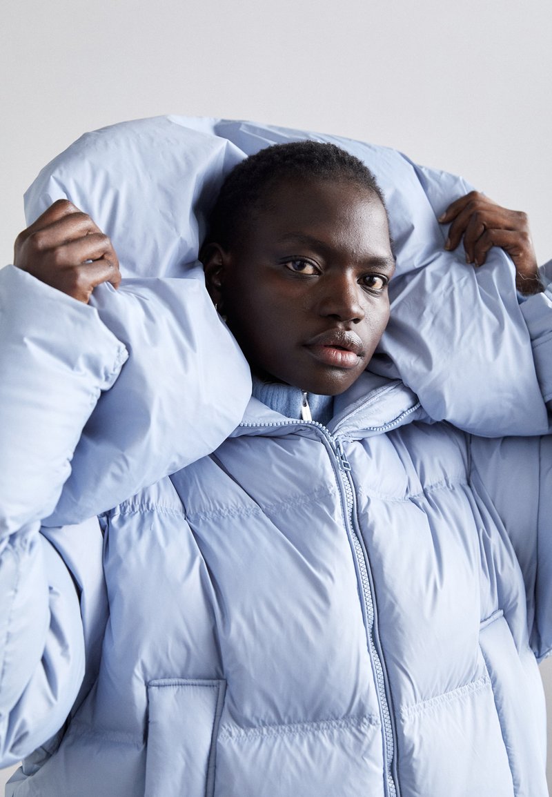 Veste doudoune bleu clair avec une texture épaisse et matelassée, capuche surdimensionnée, fermeture éclair à l'avant et poches latérales.