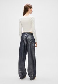 Balta ilga rankovėmis viršutinė dalis, derinama su aukštu liemeniu, plačiomis metalinėmis džinsinėmis kelnėmis, kurios turi kišenes gale ir juosmens srityje yra juodas prekės ženklas.