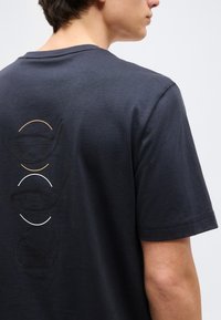 Noor inimene kannab tumedat halli T-särki, mille seljal on vertikaalselt joondatud kolm golfikepi pea disaini, millest kaks on kaunistatud kaarekujunditega.