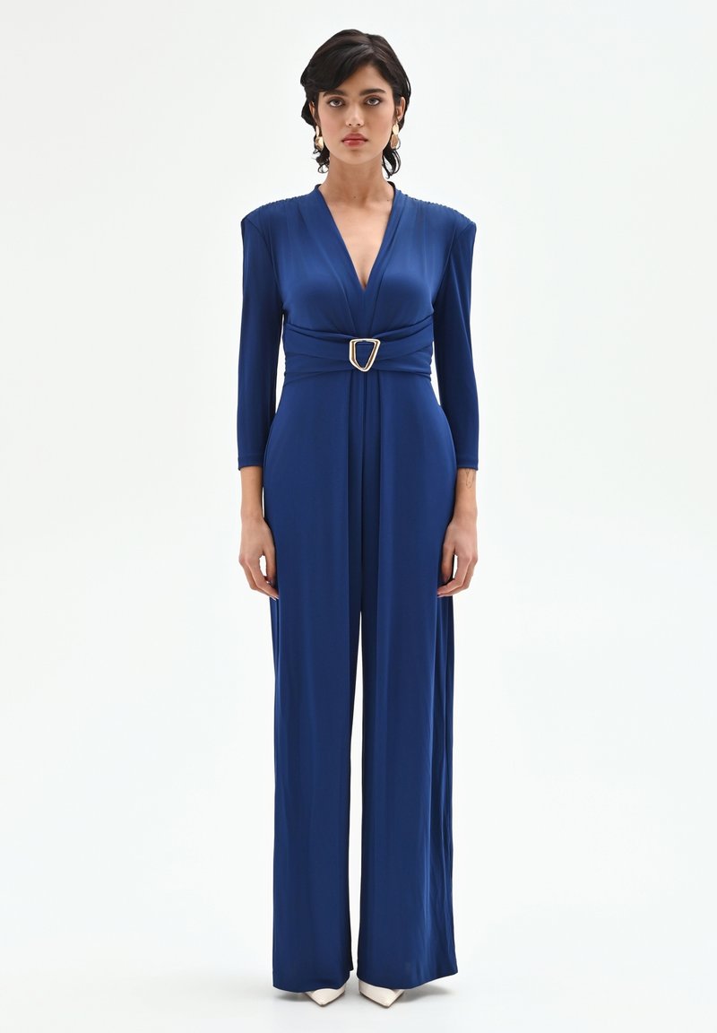 Blauer Overall mit tiefem V-Ausschnitt, langen Ärmeln, gerafften Schulterdetails und einem goldenen Schnallen-Gürtel, der die Taille betont. Glatter Stoff, weite Beine.
