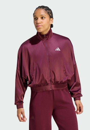 Person mit geflochtenem Haar trägt eine weinrote Jacke mit Reißverschluss und Hose mit Adidas-Logos, steht vor einem schlichten hellen Hintergrund.