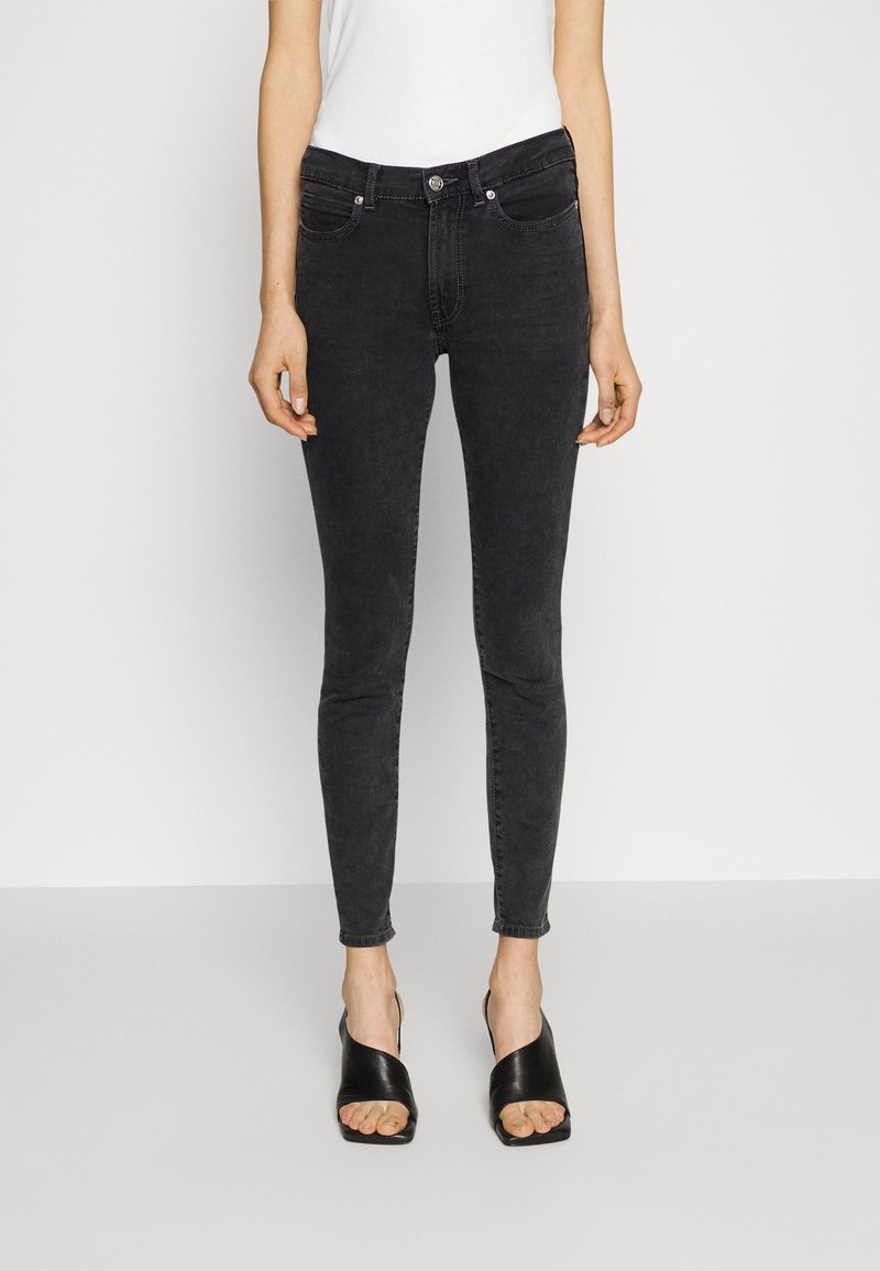 Femme portant un jean skinny taille haute noir, un haut blanc sans manches et des sandales à talons ouvertes noires, debout devant un fond uni.