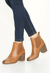 Botines de cuero marrón claro con paneles elásticos en los laterales y un tacón de bloque marrón texturizado. Combinados con jeans ajustados de color azul oscuro.