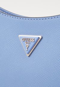 Saco de material texturizado azul com um logótipo triangular dourado e azul claro, exibindo "GUESS" e um detalhe de ponto de interrogação.